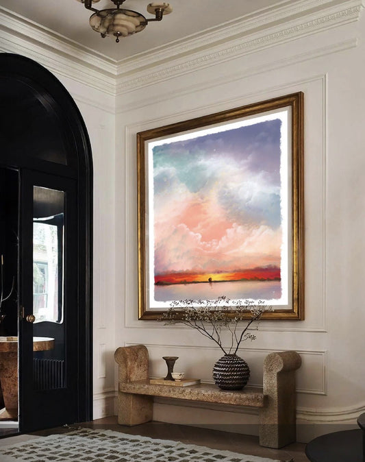 A framed art print depicting a colorful sunset over water, hung on a white wall in a styled interior room with a bench and decorative vase. Artwork by Mackenzie Thorpe is sold by The Acorn Gallery in Pocklington. 