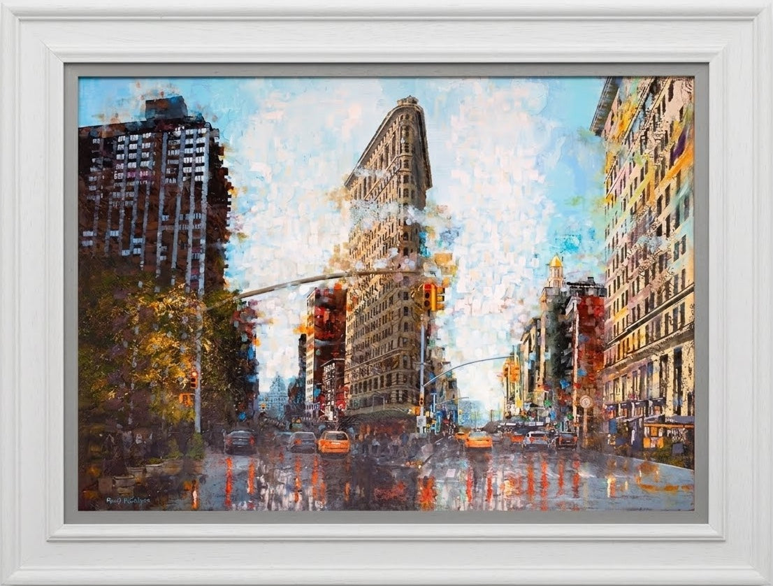 Cityscape print by Paul McIntyre featuring the Flatiron Building, warm-toned Manhattan buildings and colorful reflections on a wet street, in a white frame.