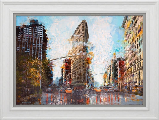 Cityscape print by Paul McIntyre featuring the Flatiron Building, warm-toned Manhattan buildings and colorful reflections on a wet street, in a white frame.