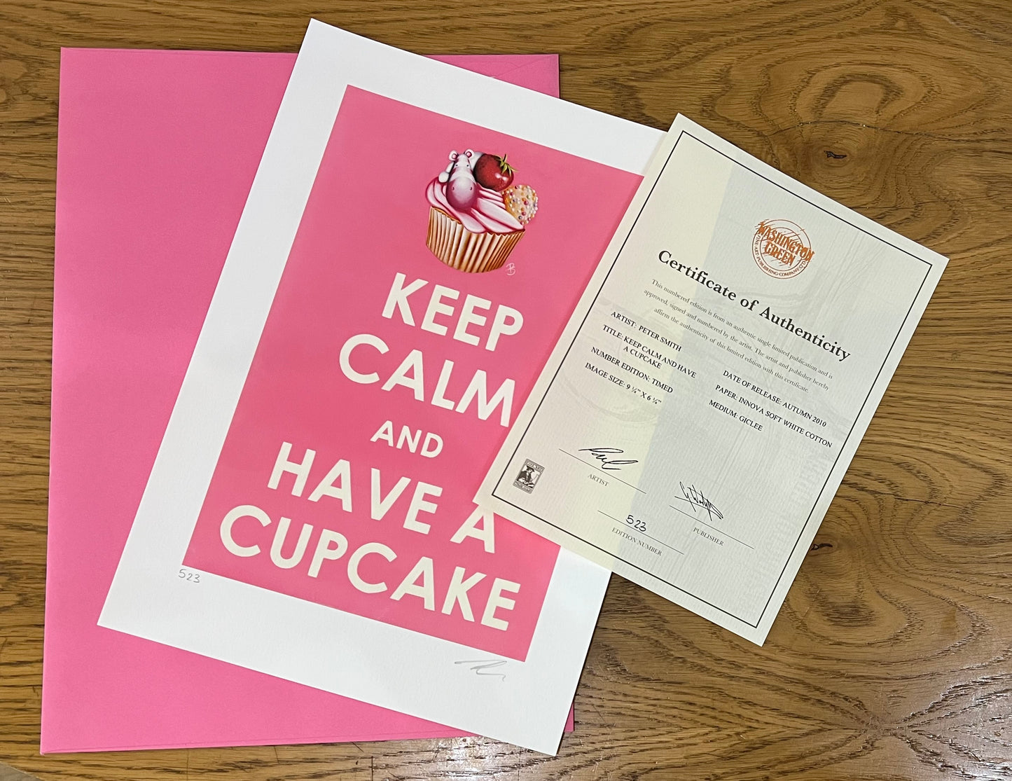 Pink and white poster with 'Keep Calm and Have a Cupcake' text and a certificate of authenticity by artist Peter Smith on a wooden surface.