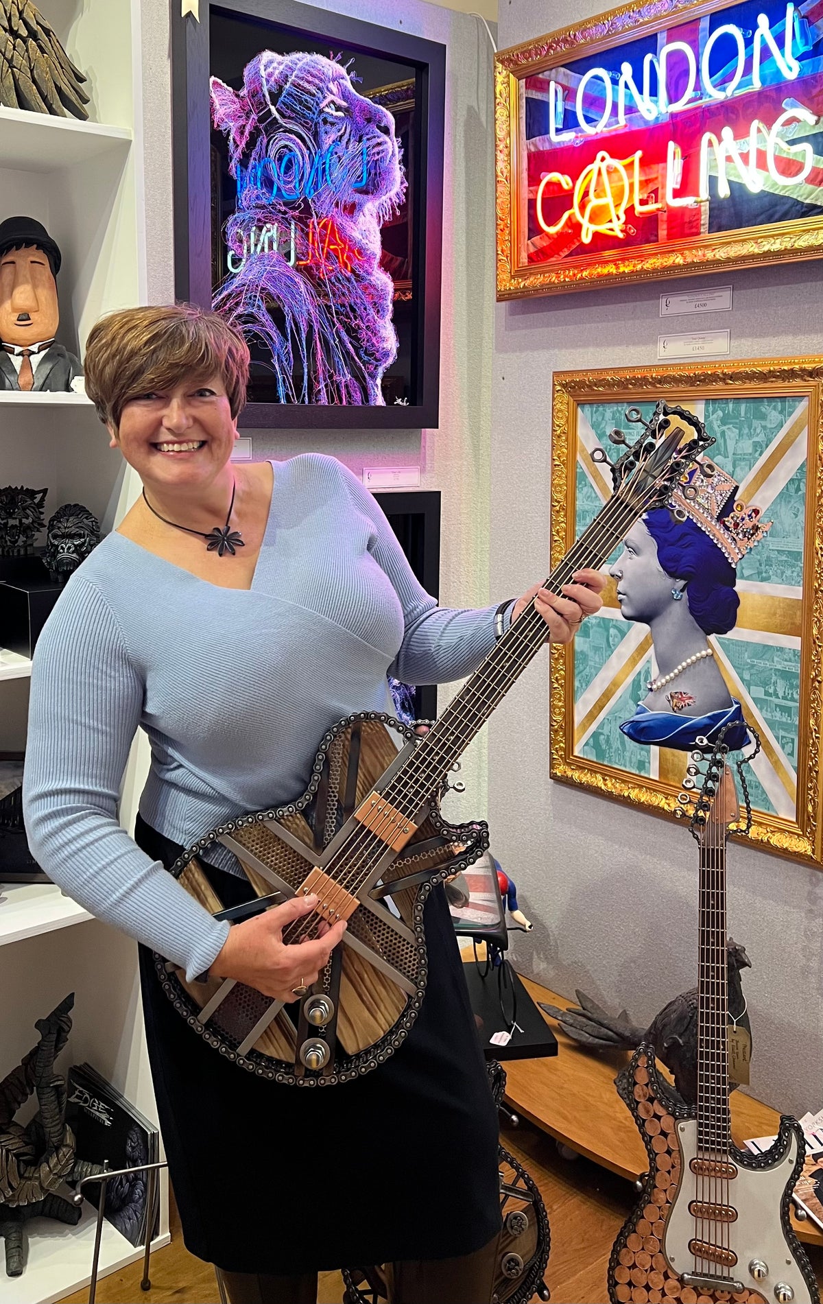 A woman in a light blue long-sleeve top and black skirt holds a decorative metal and wooden electric guitar by artist Paul Southwell in a shop interior with framed art and neon signs on the wall.