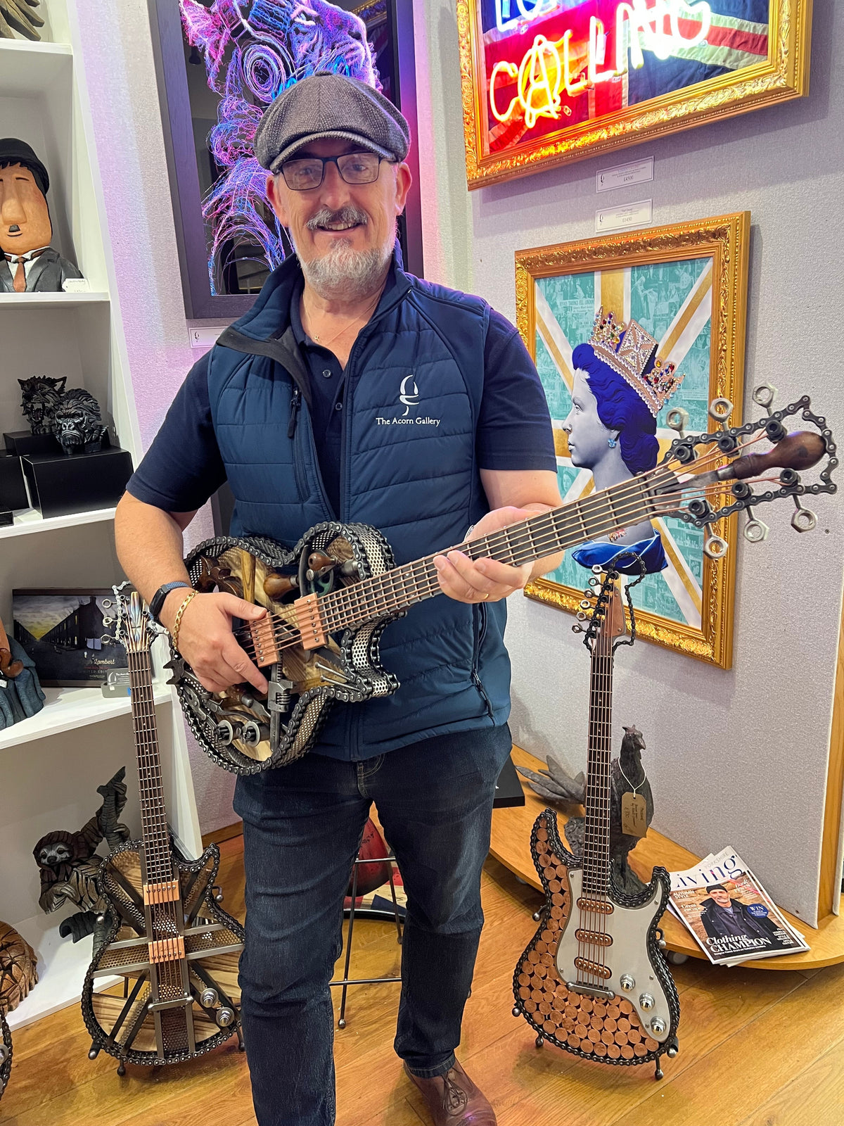 A man wearing a flat cap and navy blue puffer vest and jeans holds a unique, intricately crafted metal guitar by Paul Southwell, in a shop filled with other unusual guitars and framed art.