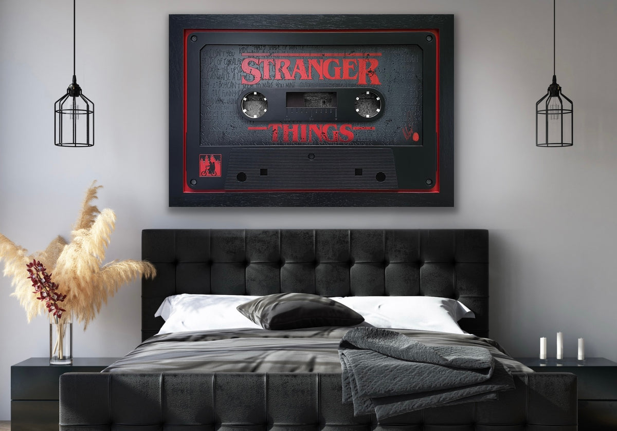 A large framed wall art piece featuring a cassette tape design with the words 'STRANGER THINGS' in red, displayed above a black tufted bed in a modern bedroom setting.