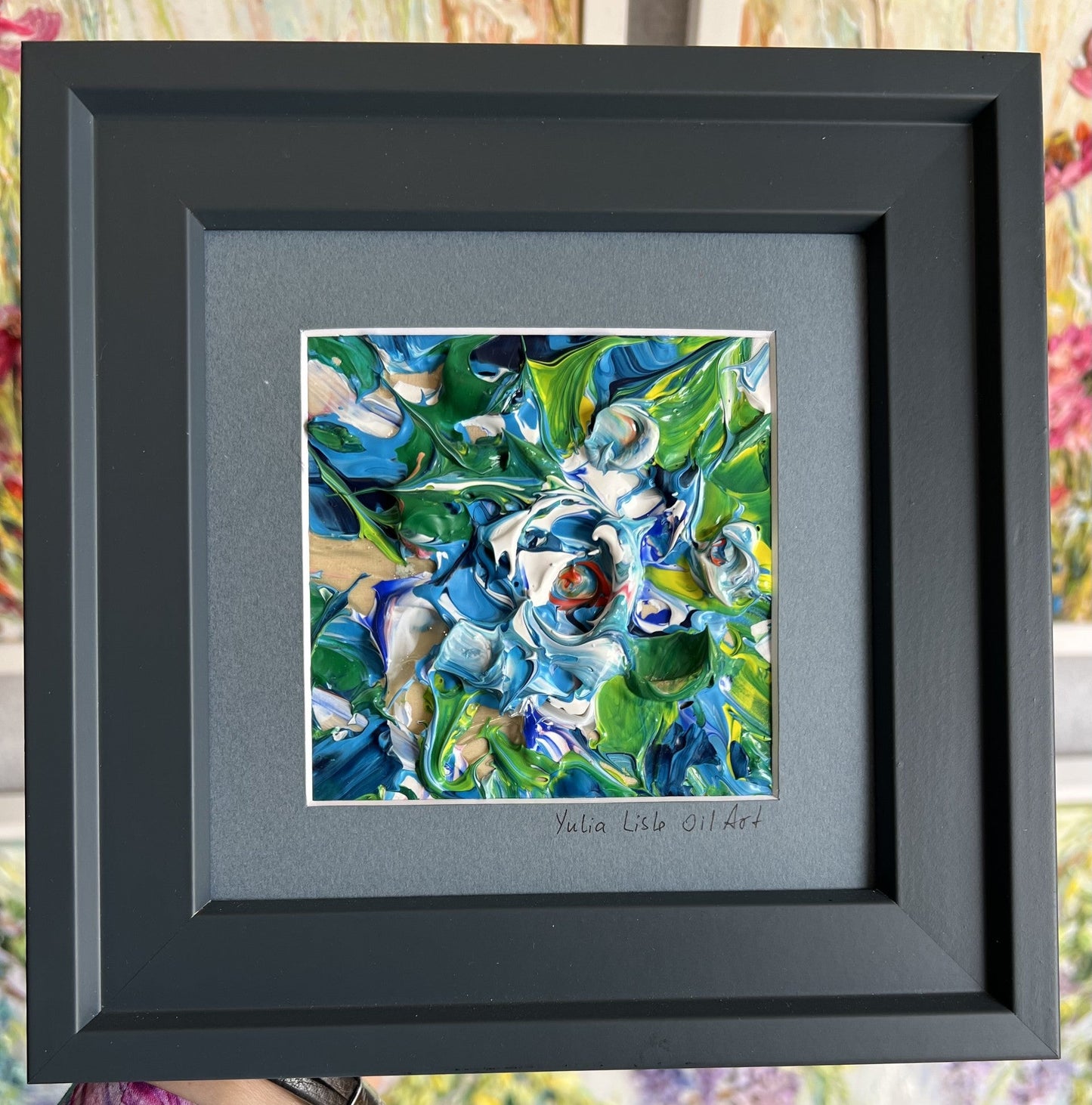 Small freestanding framed original impasto oil painting by Yulia Lisle featuring an abstract blue and white flower with vibrant green foliage, richly textured with thick palette knife strokes. Art work by Yulia Lisle and available at The Acorn Gallery in Pocklington. Shop online or call us on 01759 307652. 