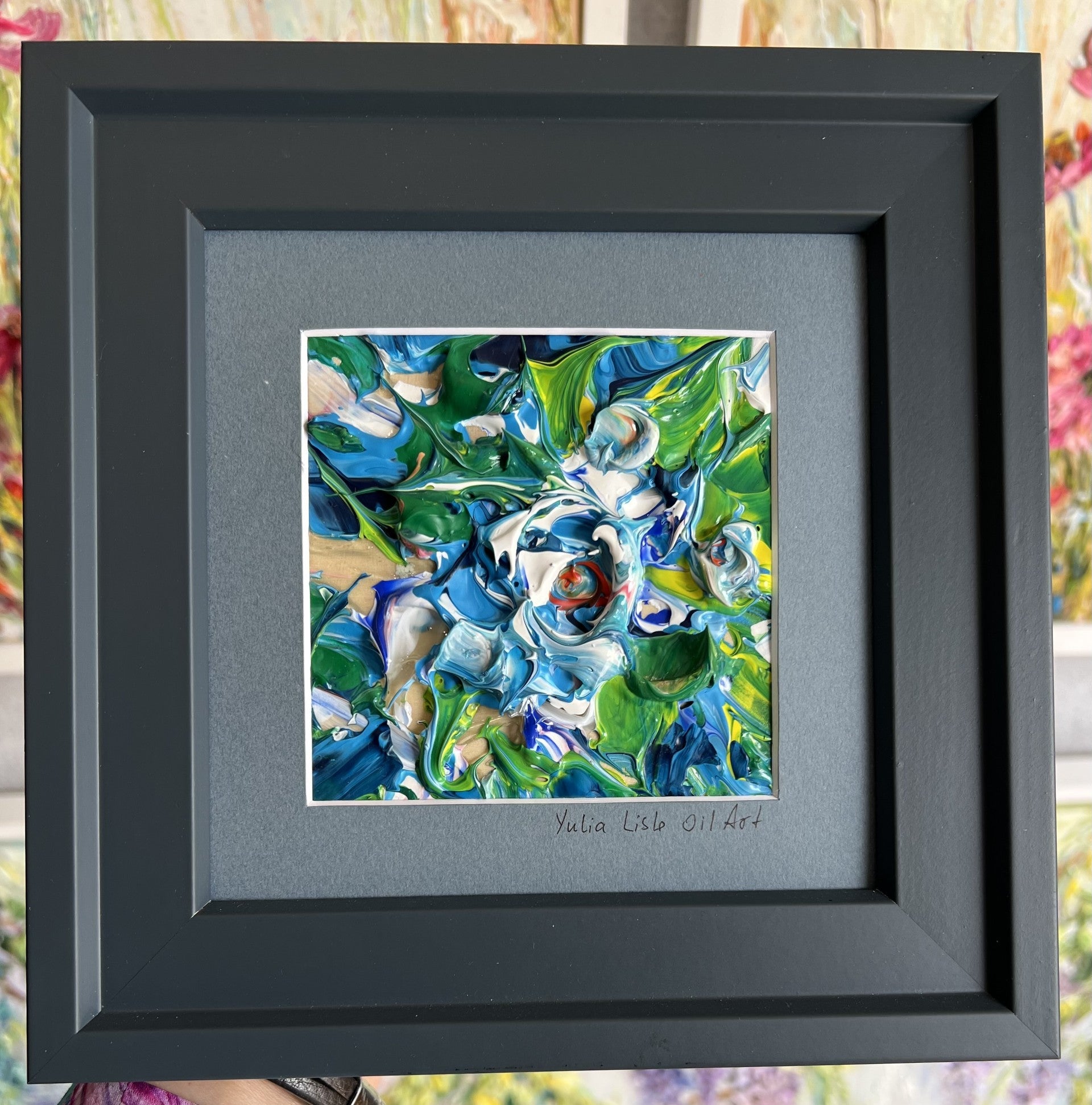 Small freestanding framed original impasto oil painting by Yulia Lisle featuring an abstract blue and white flower with vibrant green foliage, richly textured with thick palette knife strokes. Art work by Yulia Lisle and available at The Acorn Gallery in Pocklington. Shop online or call us on 01759 307652. 