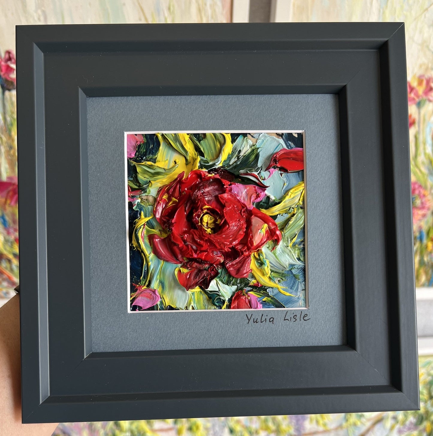 Small framed original impasto oil painting by Yulia Lisle featuring a richly textured deep red flower surrounded by vibrant green, blue and yellow foliage, created with expressive palette knife strokes. Artwork by Yulia Lisle and available at The Acor Gallery in Pocklington where you can shop online or call us on 01759 307652.
