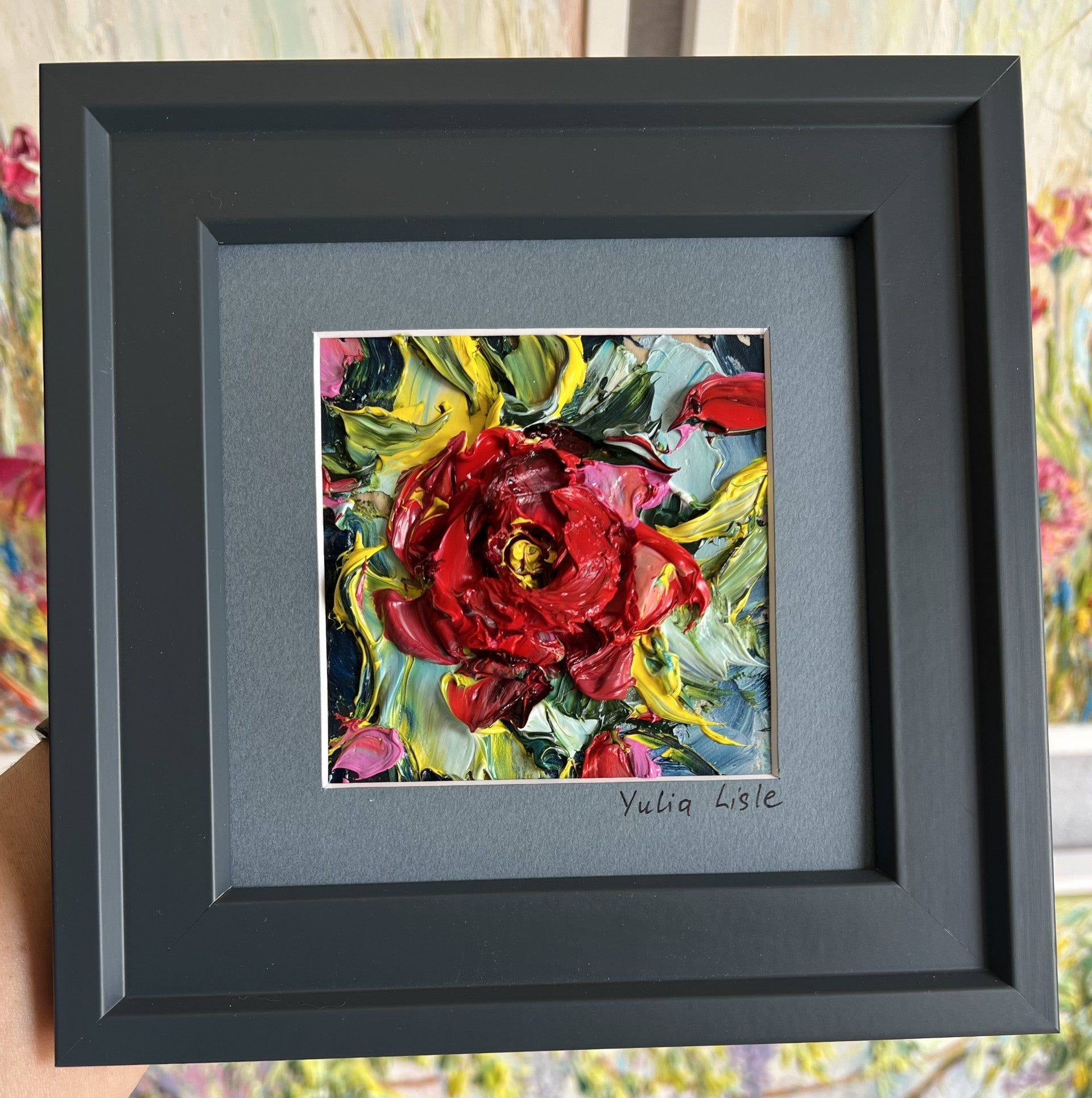 Small framed original impasto oil painting by Yulia Lisle featuring a richly textured deep red flower surrounded by vibrant green, blue and yellow foliage, created with expressive palette knife strokes. Artwork by Yulia Lisle and available at The Acor Gallery in Pocklington where you can shop online or call us on 01759 307652.