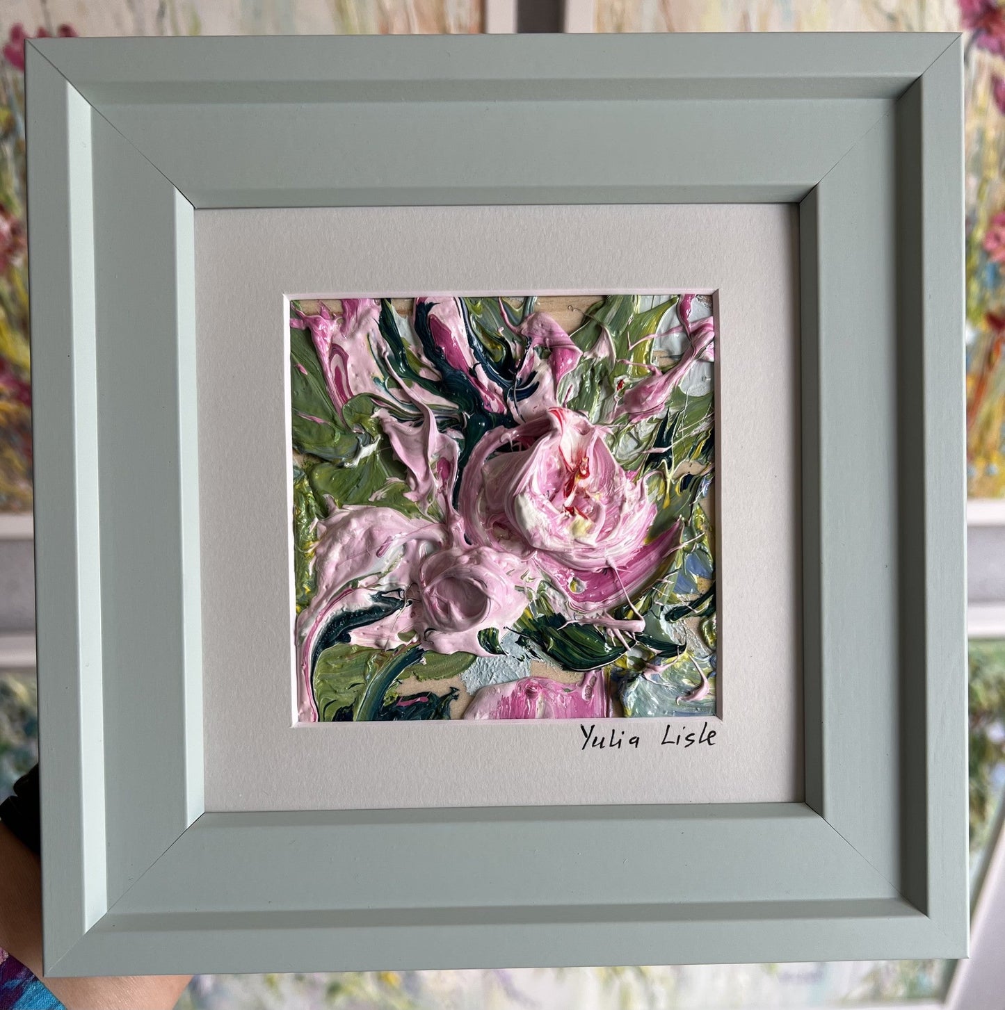 Small framed original impasto oil painting by Yulia Lisle featuring a delicately textured soft pink flower surrounded by fresh green foliage, created with expressive palette knife strokes and richly layered oil paint. Artwork by Yulia Lisle and available at The Acorn Gallery in Pocklington. Shop online or call us on 01759 307652. 